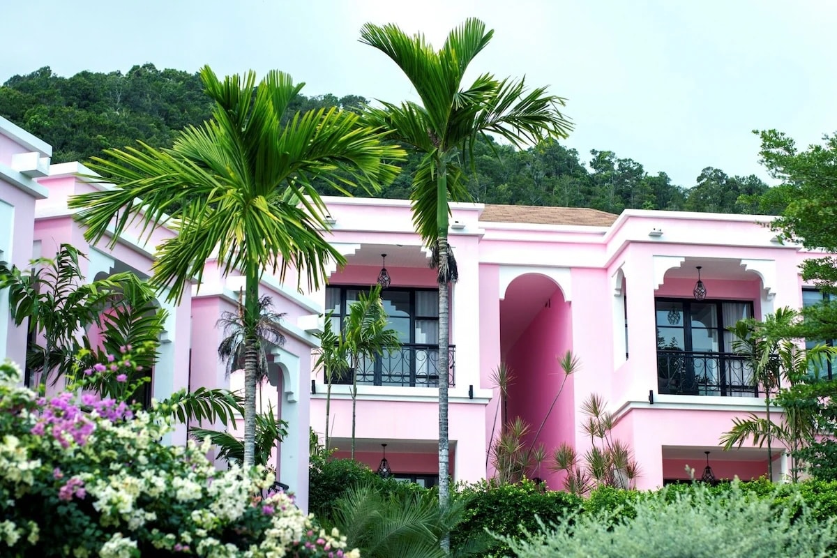 A vibrant view of Absolute Sanctuary, a vegan-friendly wellness resort in Koh Samui, Thailand. The resort features striking pink buildings with elegant arches and balconies, surrounded by lush tropical greenery and blooming flowers. The serene setting emphasizes the resort's focus on holistic health and well-being.