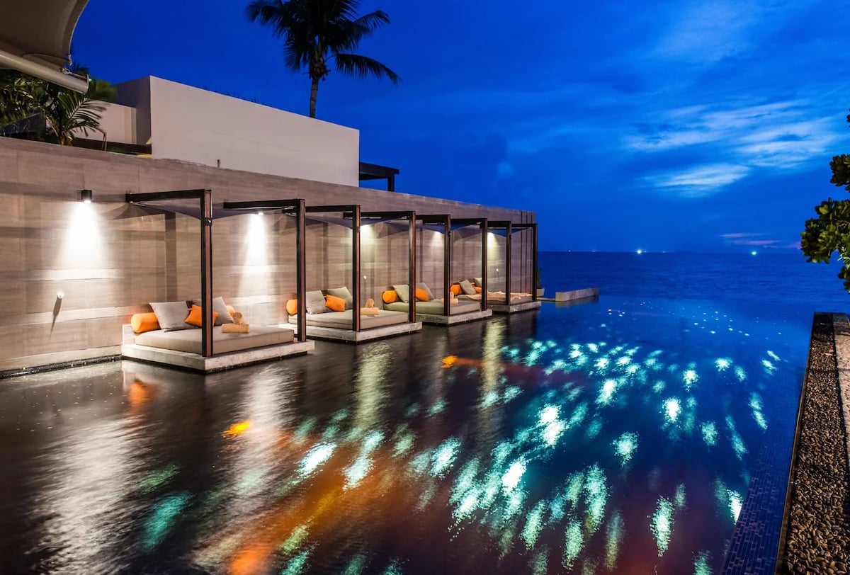 A tranquil evening scene at Aleenta, a vegan resort in Thailand. The image features a luxurious infinity pool with several poolside cabanas, each furnished with comfortable cushions and soft lighting. The pool reflects the lights and the deep blue sky, creating a serene and relaxing atmosphere by the seaside.