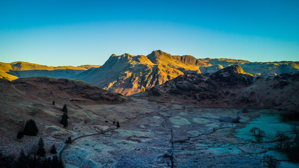 An aerial view of a mountainous landscape at sunrise, with rugged peaks and valleys bathed in golden light. The foreground shows a frosty terrain with winding paths and sparse trees, while the background highlights the majestic mountain range under a clear blue sky.