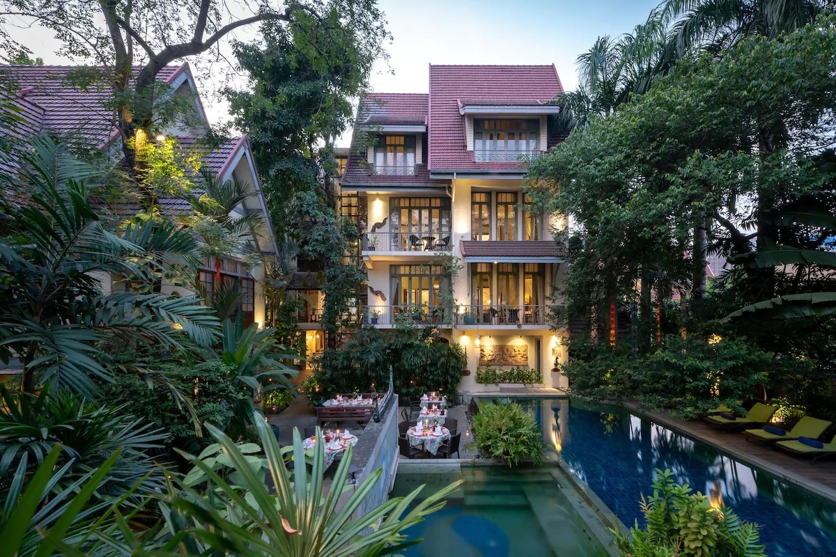 A lush and serene view of the vegan-friendly Ariyasom Villa in Bangkok, Thailand. The villa is surrounded by dense tropical vegetation and features a multi-story building with large windows and balconies. In the foreground, there's a beautifully landscaped pool area with lounge chairs and a dining area set for a meal, creating a tranquil and inviting atmosphere.