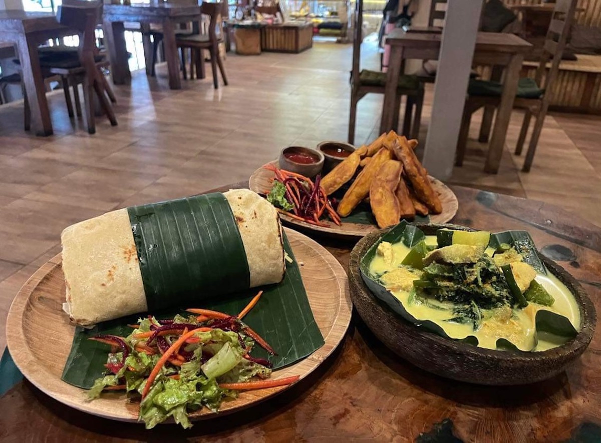A large burrito wrapped in a banana leaf, accompanied by a fresh mixed salad with carrots and leafy greens. Beside it, there are thick-cut fried plantains with dipping sauces, and a bowl of creamy yellow curry with vegetables. The food is served on wooden plates, and the setting includes wooden furniture, giving a natural, cozy atmosphere.