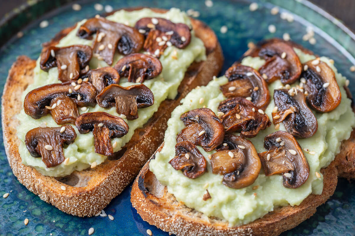 Two slices of toasted bread topped with creamy avocado spread and sautéed mushrooms, sprinkled with sesame seeds. The dish is served on a rustic blue plate, offering a fresh and savory vegan option from a Bali café.