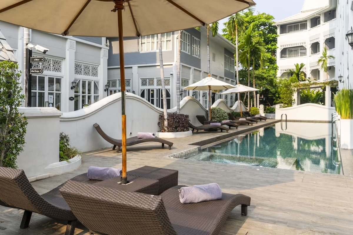 A serene poolside view at Away Chiang Mai Thapae Resort, a 100% vegan and eco-friendly resort in Chiang Mai, Thailand. The image features elegant lounge chairs with rolled towels and large umbrellas for shade, set around a pristine swimming pool. The resort's beautiful architecture, with white and gray buildings, provides a tranquil and luxurious atmosphere.