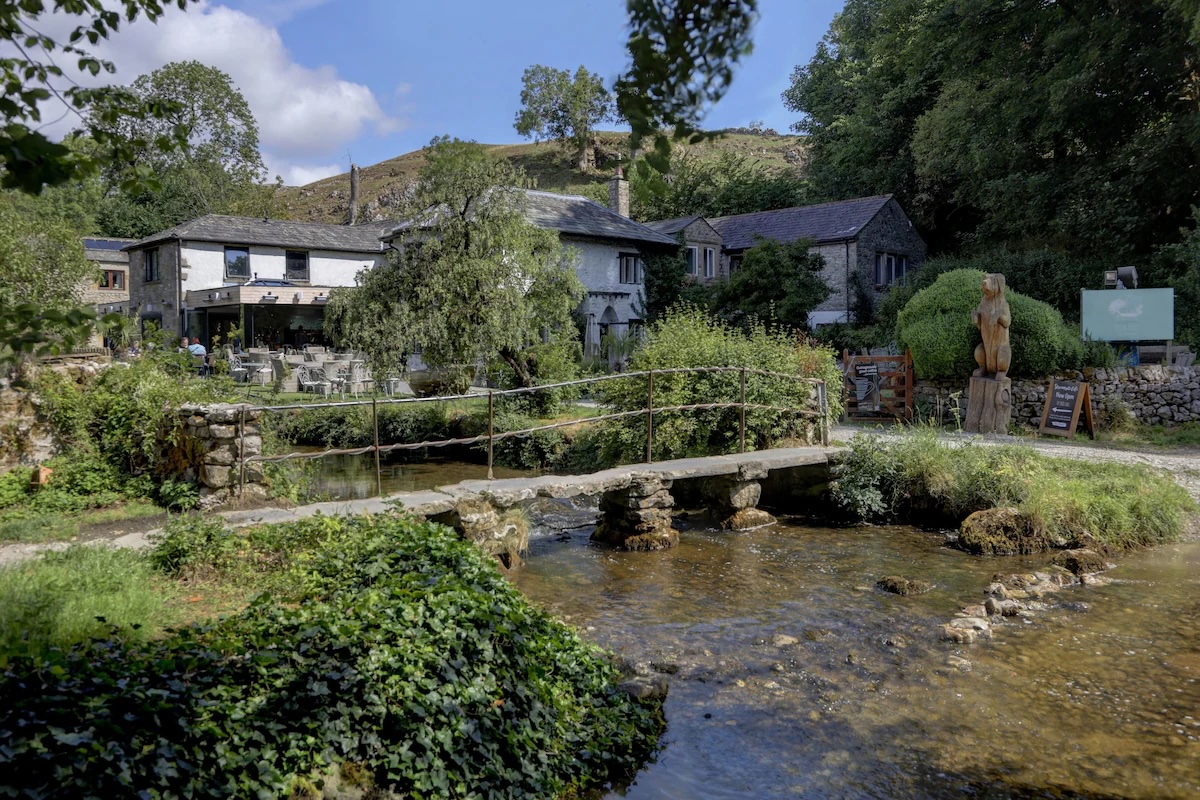 Scenic view of Beck Hall Hotel, Bar & Restaurant in Yorkshire Dales National Park, England’s first 100% plant-based hotel, surrounded by lush greenery and a tranquil stream with a stone bridge.
