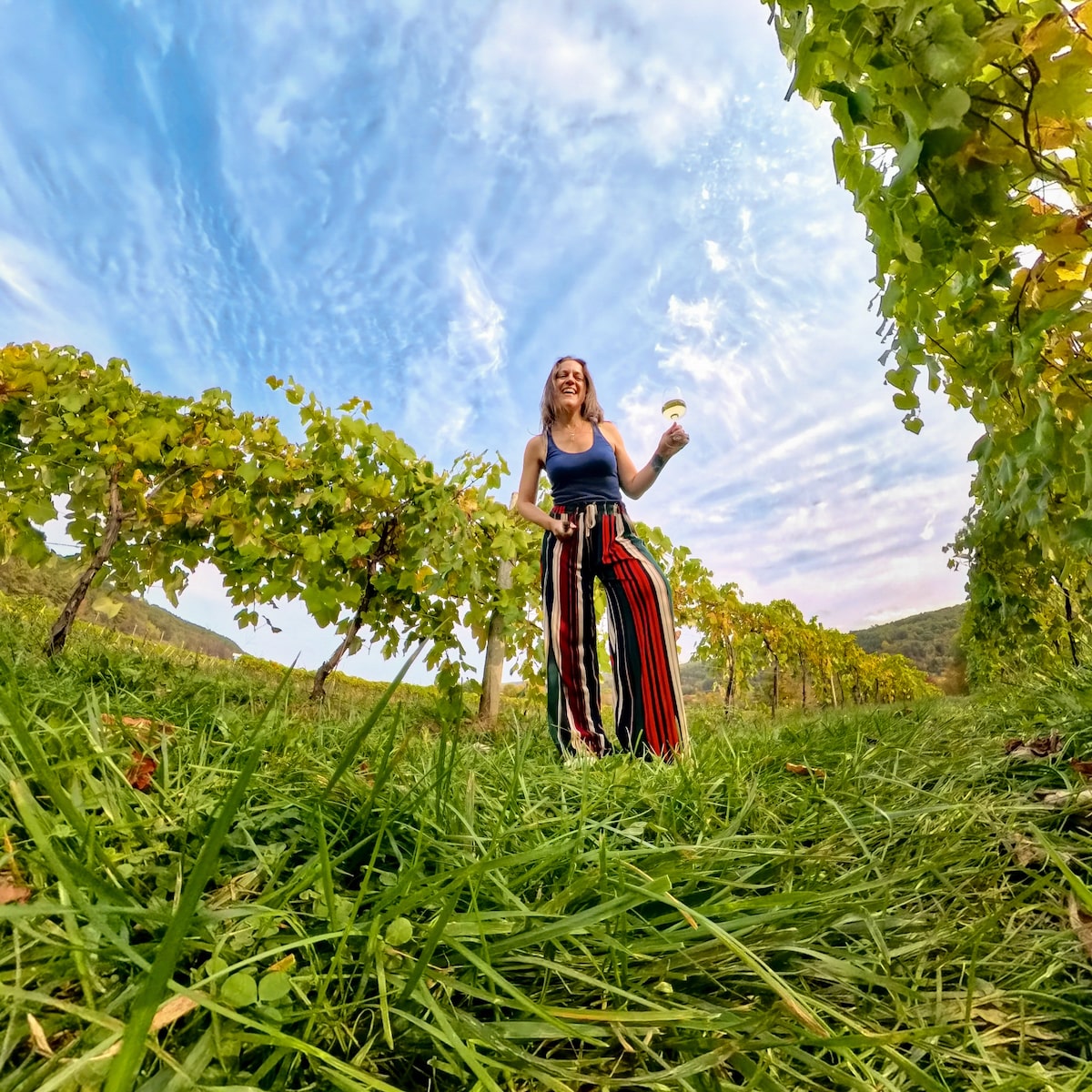 Woman enjoying a glass of wine while standing in the lush vineyard rows at Benjamin Bridge Winery in Wolfville, Nova Scotia, surrounded by grapevines and blue skies.