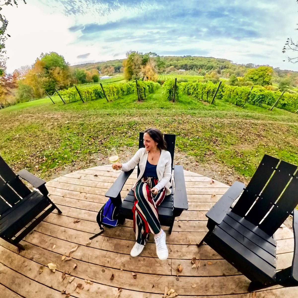 Woman enjoying a glass of white wine while relaxing in an Adirondack chair with scenic views of grapevines and rolling hills at Benjamin Bridge Vineyard in Wolfville, Nova Scotia.