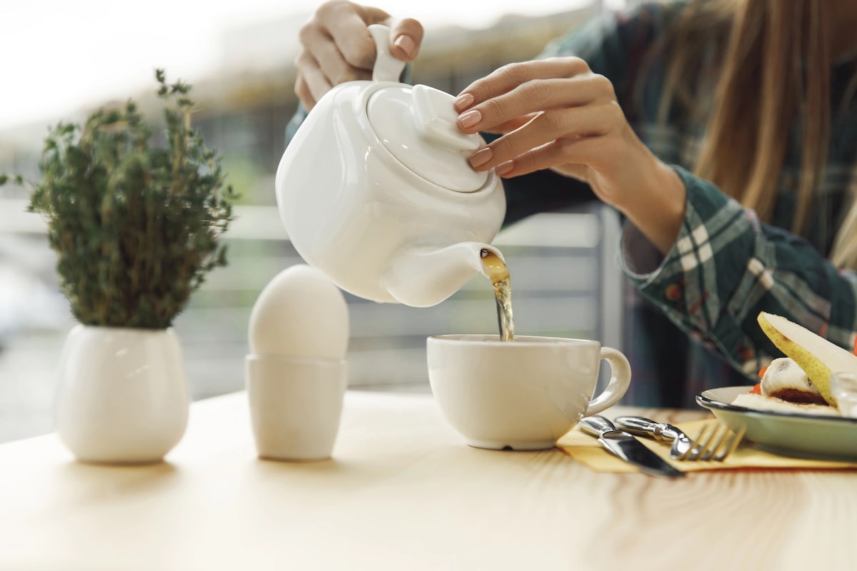 woman pouring tea into a white mug while sitting at a table and eating a vegan snack