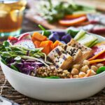 A colorful vegan Buddha bowl featuring grilled tofu topped with black sesame seeds, fresh greens, chickpeas, quinoa, purple sweet potatoes, vibrant orange slices, and assorted vegetables like cucumbers and shredded cabbage. The bowl is accompanied by a glass of iced tea in the background, offering a wholesome and nourishing plant-based meal.