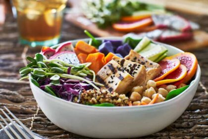 A colorful vegan Buddha bowl featuring grilled tofu topped with black sesame seeds, fresh greens, chickpeas, quinoa, purple sweet potatoes, vibrant orange slices, and assorted vegetables like cucumbers and shredded cabbage. The bowl is accompanied by a glass of iced tea in the background, offering a wholesome and nourishing plant-based meal.