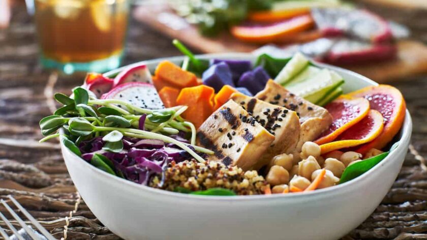 A colorful vegan Buddha bowl featuring grilled tofu topped with black sesame seeds, fresh greens, chickpeas, quinoa, purple sweet potatoes, vibrant orange slices, and assorted vegetables like cucumbers and shredded cabbage. The bowl is accompanied by a glass of iced tea in the background, offering a wholesome and nourishing plant-based meal.