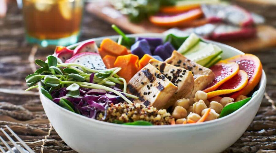 A colorful vegan Buddha bowl featuring grilled tofu topped with black sesame seeds, fresh greens, chickpeas, quinoa, purple sweet potatoes, vibrant orange slices, and assorted vegetables like cucumbers and shredded cabbage. The bowl is accompanied by a glass of iced tea in the background, offering a wholesome and nourishing plant-based meal.