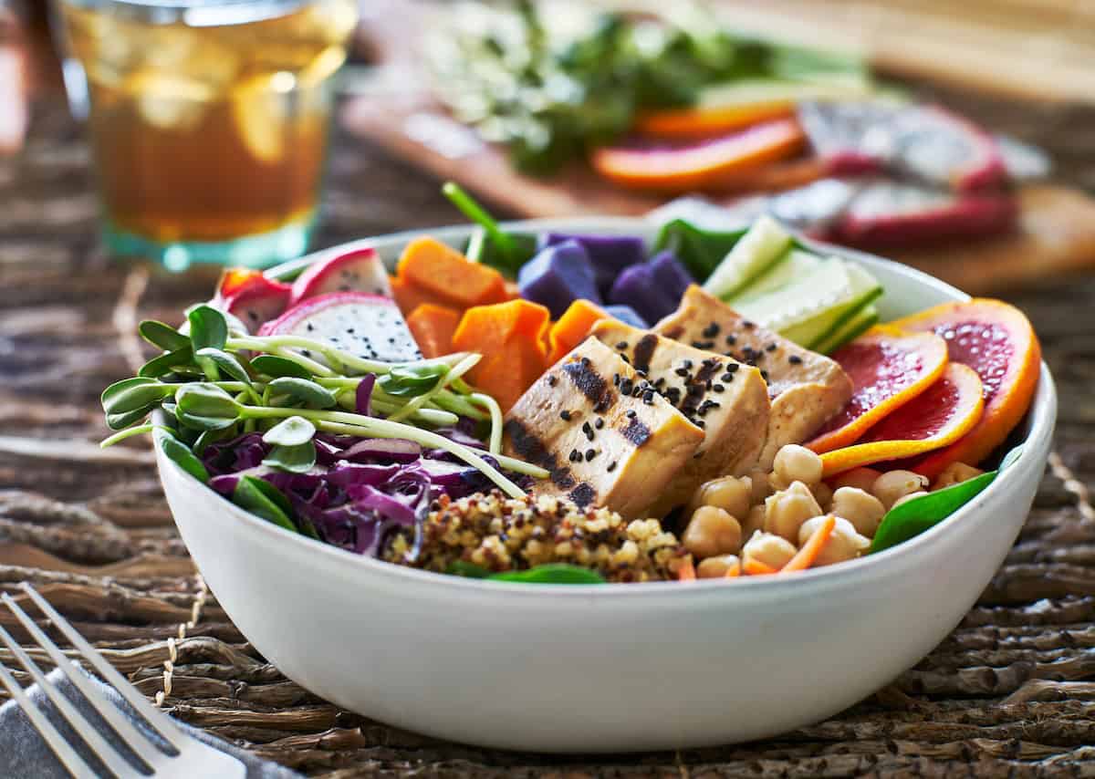 A colorful vegan Buddha bowl featuring grilled tofu topped with black sesame seeds, fresh greens, chickpeas, quinoa, purple sweet potatoes, vibrant orange slices, and assorted vegetables like cucumbers and shredded cabbage. The bowl is accompanied by a glass of iced tea in the background, offering a wholesome and nourishing plant-based meal.