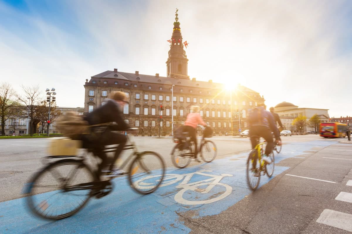 Cyclists riding along a dedicated bike lane in Copenhagen, Denmark, with the historic Christiansborg Palace in the background and the sun shining through the sky, highlighting the city's bike-friendly culture.