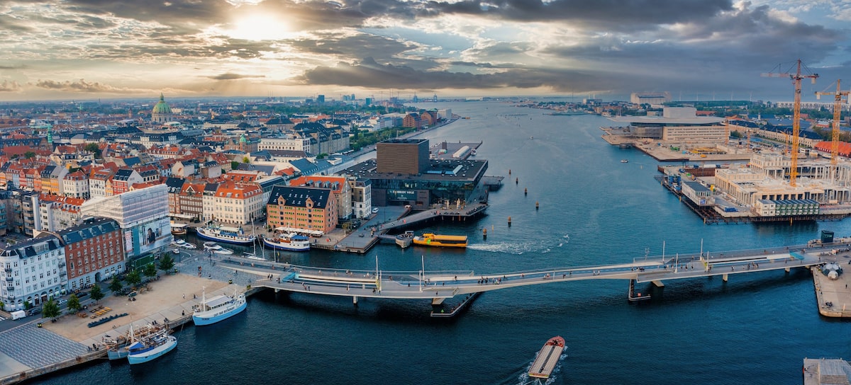 Aerial view of Copenhagen’s waterfront at sunset, featuring colorful historic buildings, modern architecture, a pedestrian bridge, and boats navigating the harbor. The city skyline blends tradition with contemporary development.