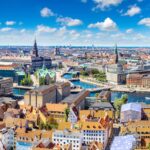 A panoramic view of Copenhagen, Denmark, showcasing colorful rooftops, historic buildings, and the city's scenic canals under a bright blue sky with fluffy white clouds.