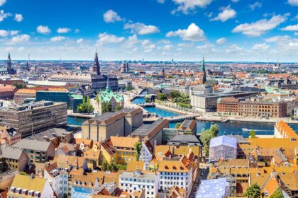 A panoramic view of Copenhagen, Denmark, showcasing colorful rooftops, historic buildings, and the city's scenic canals under a bright blue sky with fluffy white clouds.