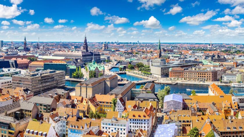 A panoramic view of Copenhagen, Denmark, showcasing colorful rooftops, historic buildings, and the city's scenic canals under a bright blue sky with fluffy white clouds.