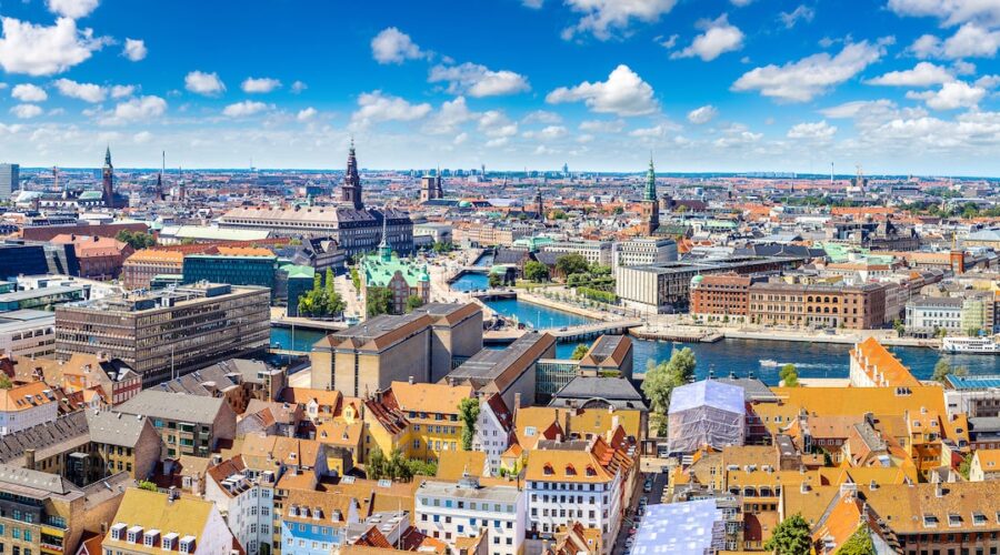 A panoramic view of Copenhagen, Denmark, showcasing colorful rooftops, historic buildings, and the city's scenic canals under a bright blue sky with fluffy white clouds.