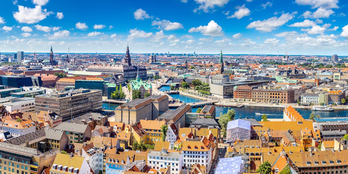A panoramic view of Copenhagen, Denmark, showcasing colorful rooftops, historic buildings, and the city's scenic canals under a bright blue sky with fluffy white clouds.