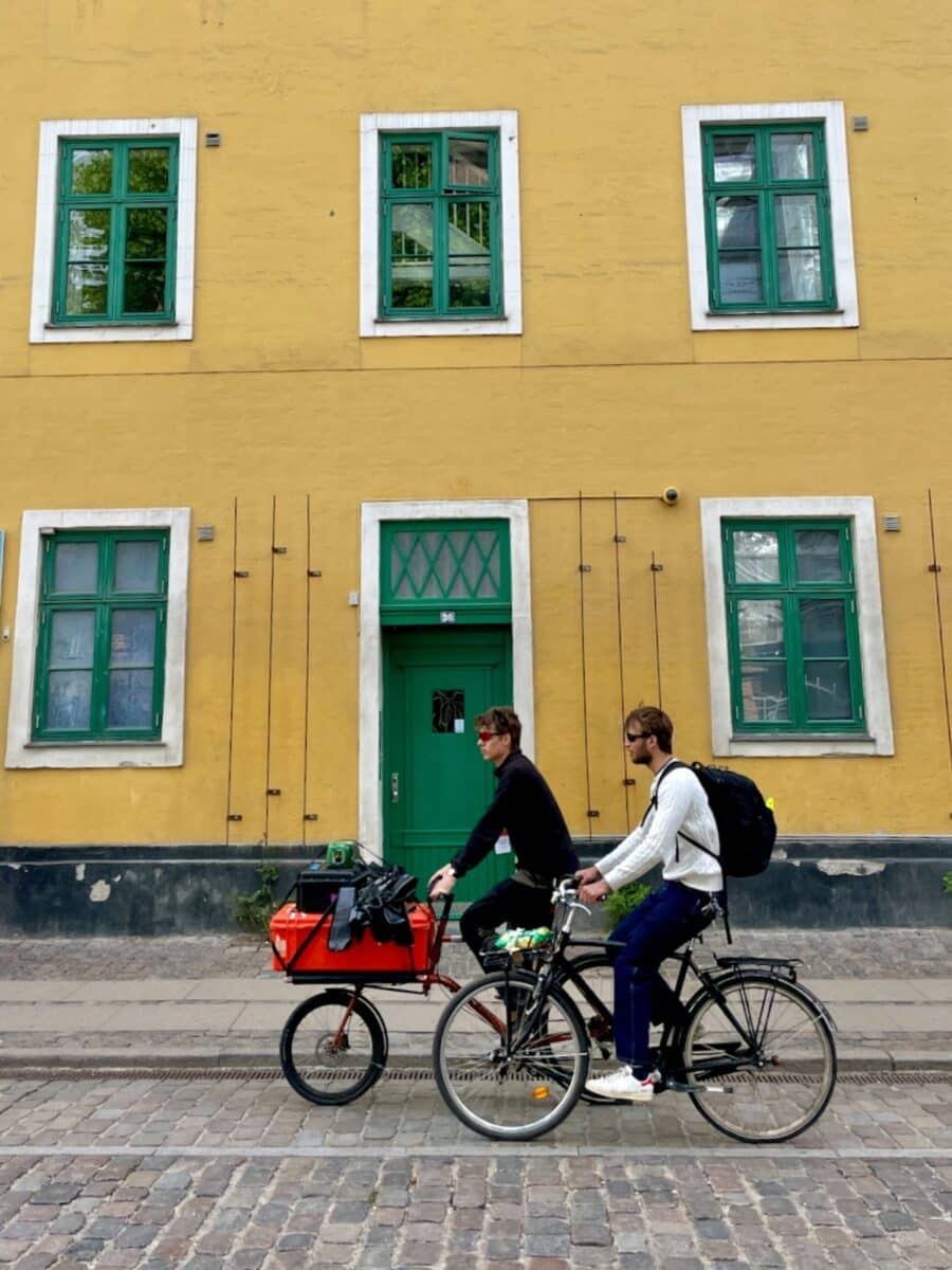 Two guys cycling on cobblestone streets past yellow buildings in Copenhagen, Denmark