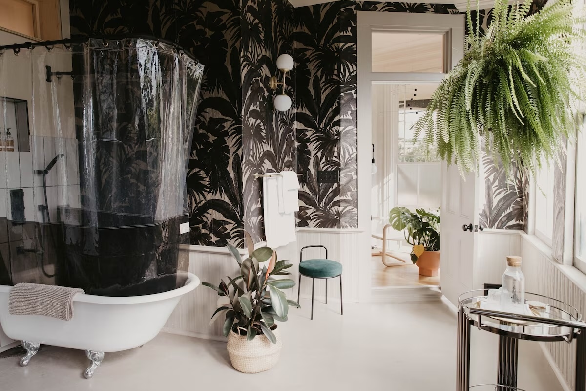 Chic and modern bathroom at the vegan boutique hotel Dreamers by Dreamers Welcome in Wilmington, North Carolina, featuring a clawfoot tub with a clear shower curtain, lush plants, and tropical-themed wallpaper.