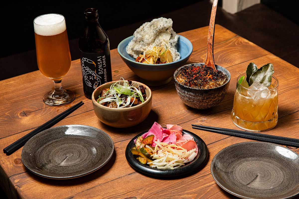 A table set with a selection of vegan East Asian small plates, pickled vegetables, rice dishes, and drinks at Bonsai Plant Kitchen in Brighton, including craft beer and a cocktail garnished with kaffir lime leaves.