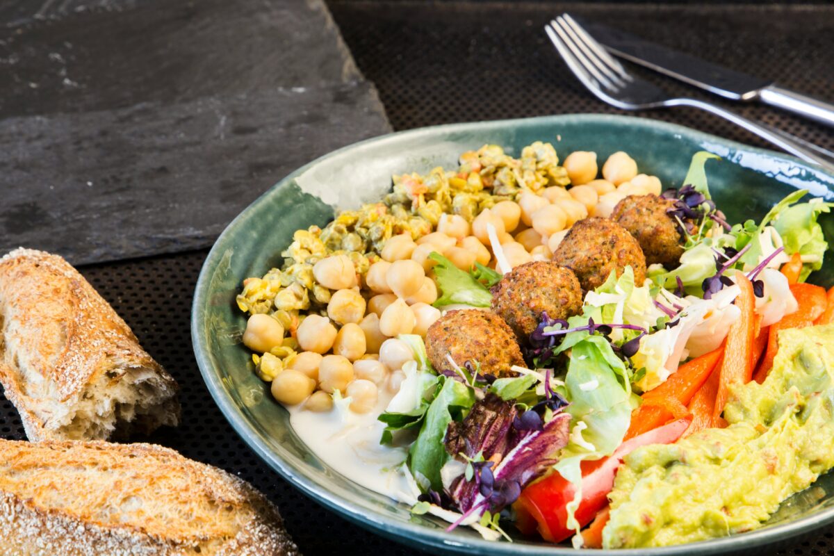 A falafel platter served in a green bowl. The dish features golden-brown falafel balls, chickpeas, fresh mixed greens, red bell pepper slices, and a scoop of guacamole, accompanied by a creamy dressing. On the side, there is a portion of crusty bread.
