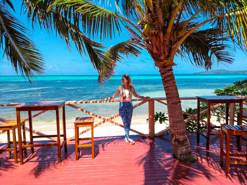 A girl standing on a pink terrace under a palm tree with the striped blue sea of the lagoon in the Yasawas behind. 