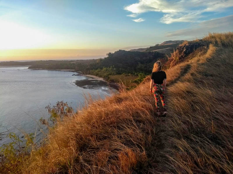 A girl walking on a grassy ridge overlooking a beach at sunset in Fiji. Hiking is a good way to experience ecotourism in Fiji. 