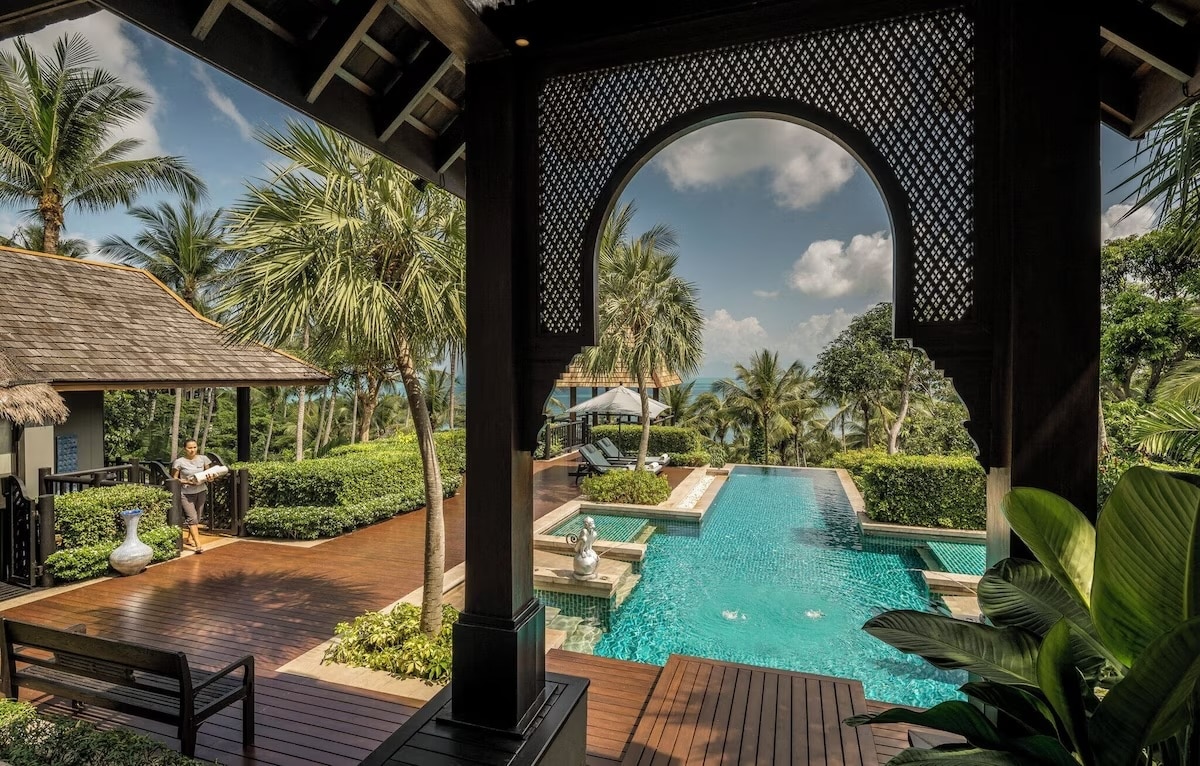 A picturesque view of the luxurious eco-friendly Four Seasons Resort in Koh Samui, Thailand. The image features a beautifully designed infinity pool surrounded by lush tropical greenery and elegant wooden decking. A traditional Thai-style pavilion frames the scene, offering a serene and opulent atmosphere with views of palm trees and the ocean in the background.