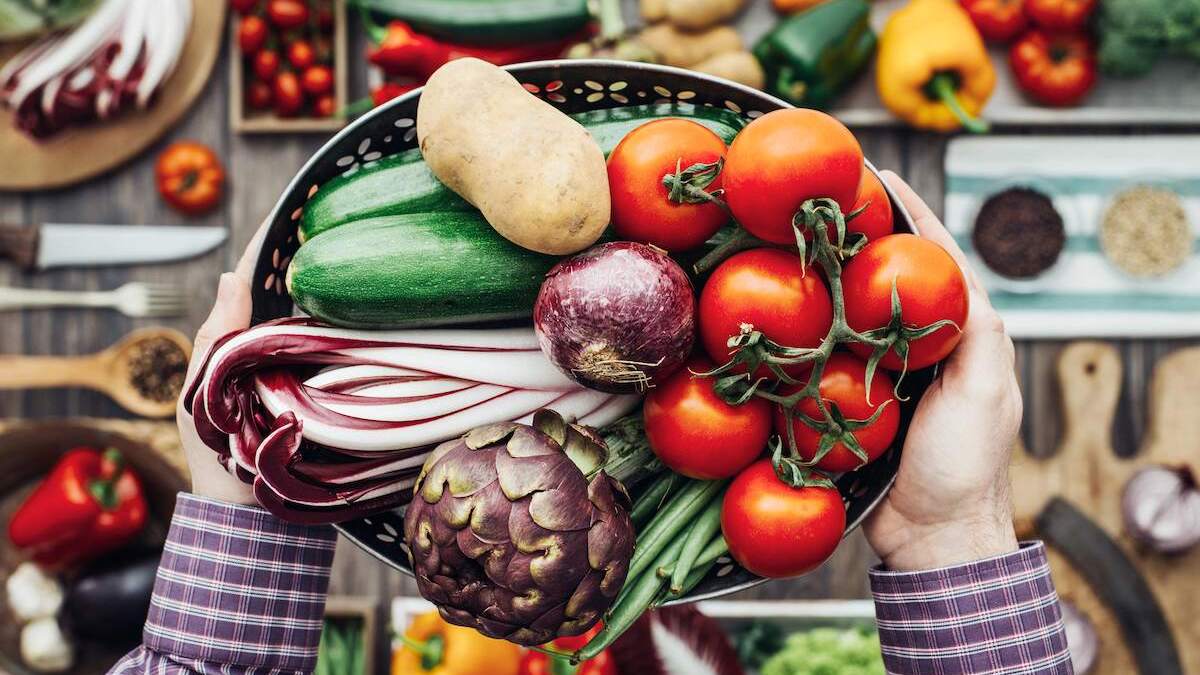 Hands holding a basket filled with fresh vegetables, including tomatoes, zucchini, red onion, radicchio, and an artichoke.