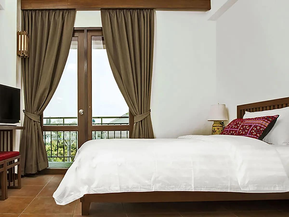 A serene and inviting room at Green Tiger House, Chiang Mai's first plant-based hotel. The room features a comfortable bed with white linens, accented by a colorful pillow, and a small wooden desk with a chair. Large windows with brown curtains allow natural light to fill the space, highlighting the hotel's focus on fresh, organic ingredients and a tranquil atmosphere.