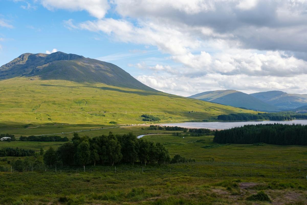 A vast, lush green landscape with rolling hills and a mountain in the background under a partly cloudy sky. A winding river flows through the valley, and dense forests can be seen in the distance.