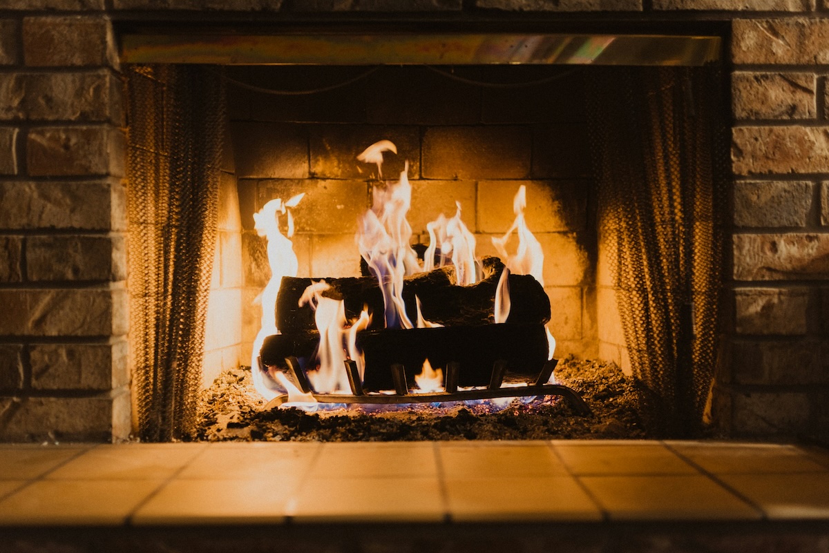 A cozy brick fireplace with a roaring fire, providing warmth and ambiance. The flames dance around the logs, casting a soft glow on the surrounding brick and tile hearth.
