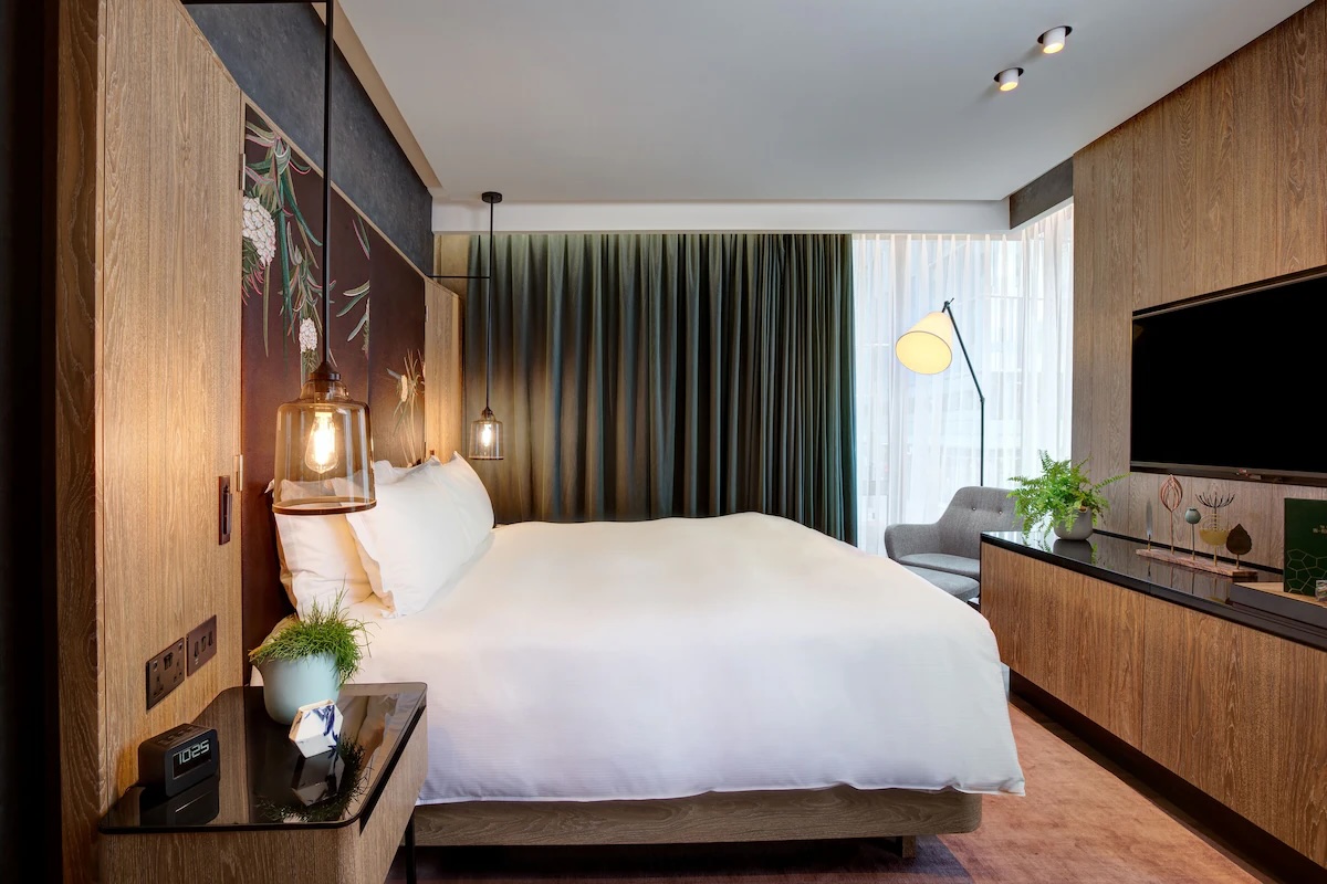 A modern hotel room at the vegan Hilton London Bankside in the UK featuring a large, comfortable bed with white linens, stylish hanging lamps, and wooden furnishings. The room includes a seating area with a chair and floor lamp, a large window with heavy curtains, and a sleek television mounted on the wall.