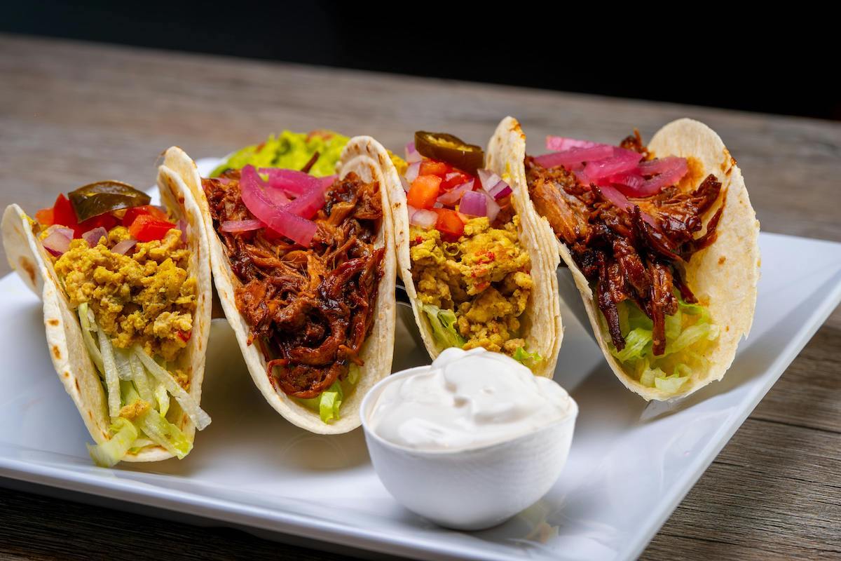 A plate of vegan tacos. Two of the tacos are filled with shredded jackfruit, and topped with pickled red onions. The other tacos contain a tofu scramble or similar plant-based protein, garnished with jalapeños, diced tomatoes, and lettuce. A small bowl of creamy vegan sour cream is placed on the side of the plate for dipping.