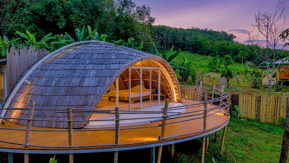 An eco-friendly resort at Jaiyen Eco Resort in Koh Yao Noi, Thailand. The image showcases a unique, dome-shaped bamboo bungalow with large glass doors, nestled in a lush green environment. The bungalow is elevated on stilts with a wraparound wooden deck, blending seamlessly with the surrounding natural landscape, emphasizing the resort's commitment to sustainability and eco-friendly practices.