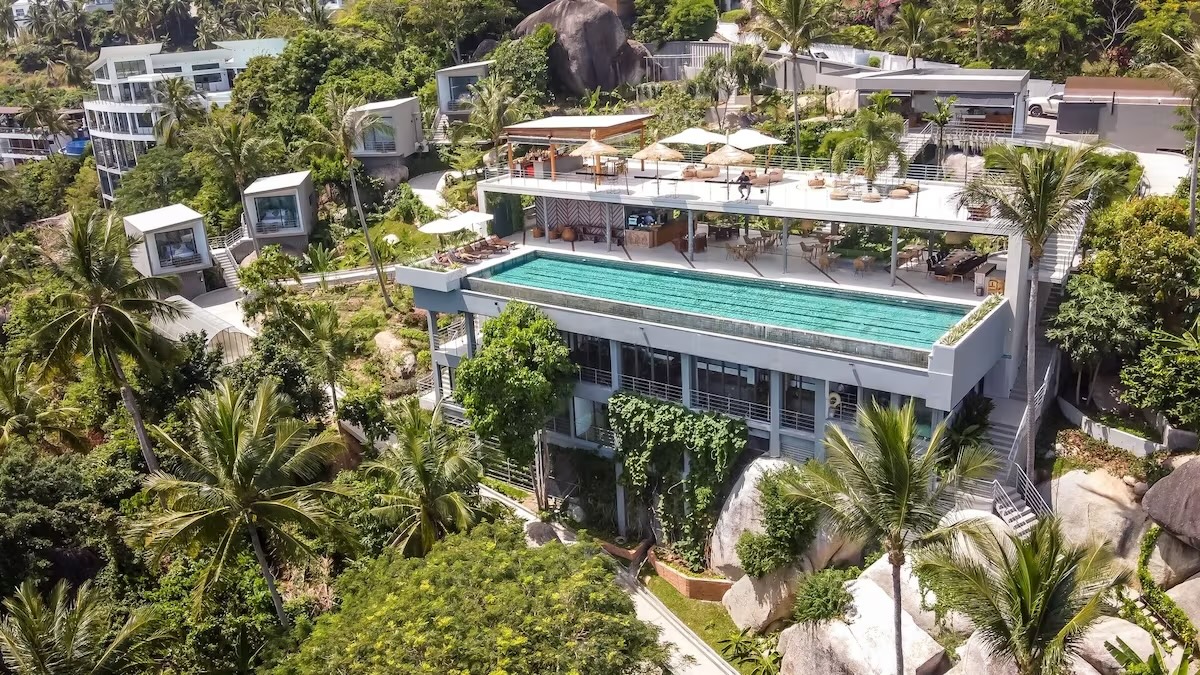 An aerial view of Kapuhala Koh Samui Boutique Wellness Resort, a vegan resort in Koh Samui, Thailand, near Chaweng Beach. The resort features modern architecture with several sleek buildings nestled among lush tropical greenery. A striking infinity pool and a spacious terrace with lounge areas and umbrellas are prominently visible, offering stunning views of the surrounding landscape.