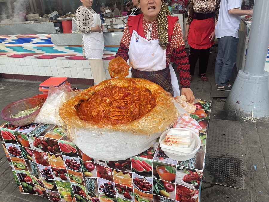 A vendor at Chorsu Bazaar in Uzbekistan standing behind a large bowl of Uzbek 'lasagna', a layered vegetable and noodle dish. This vegetarian-friendly option showcases the diverse and delicious vegetarian dishes available in Uzbek cuisine.