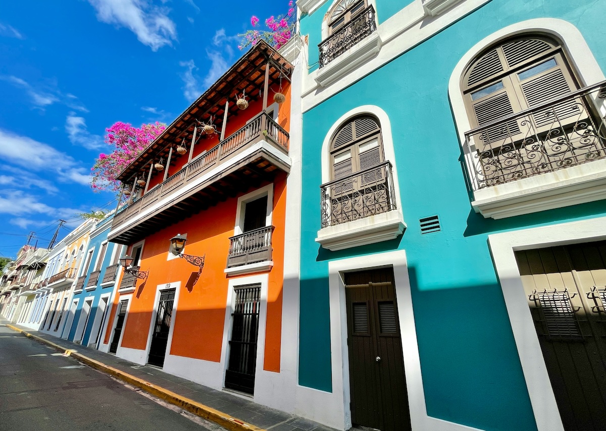 Vibrant, colorful buildings with ornate balconies line a narrow street in San Juan, Puerto Rico, against a backdrop of a clear blue sky. The bright orange and teal facades are adorned with flowering plants, reflecting the city's lively culture and architecture.
