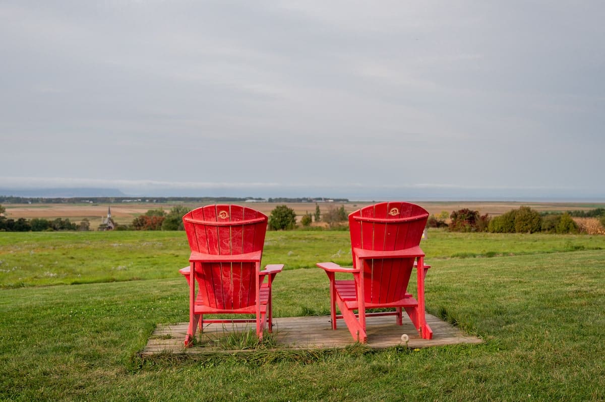 Two red Adirondack chairs overlooking the scenic landscape of Grand Pré View Park, with expansive views of the Annapolis Valley, accessible via a trail at Domaine de Grand Pré
