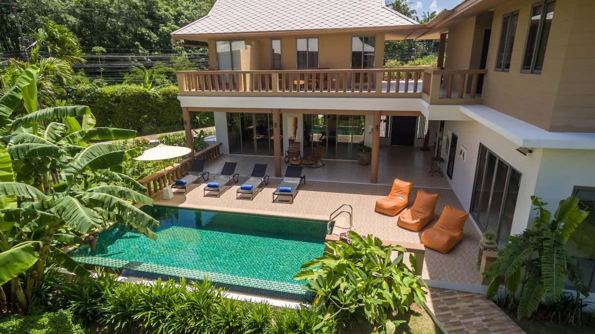 An inviting view of TheLifeCo Wellbeing and Detox Center, an all-inclusive vegan-friendly resort in Phuket, Thailand. The image features a spacious two-story villa with a wraparound balcony, lounge chairs, and bean bags arranged around a pristine swimming pool. The surrounding area is lush with tropical plants, contributing to the resort's tranquil and rejuvenating atmosphere.