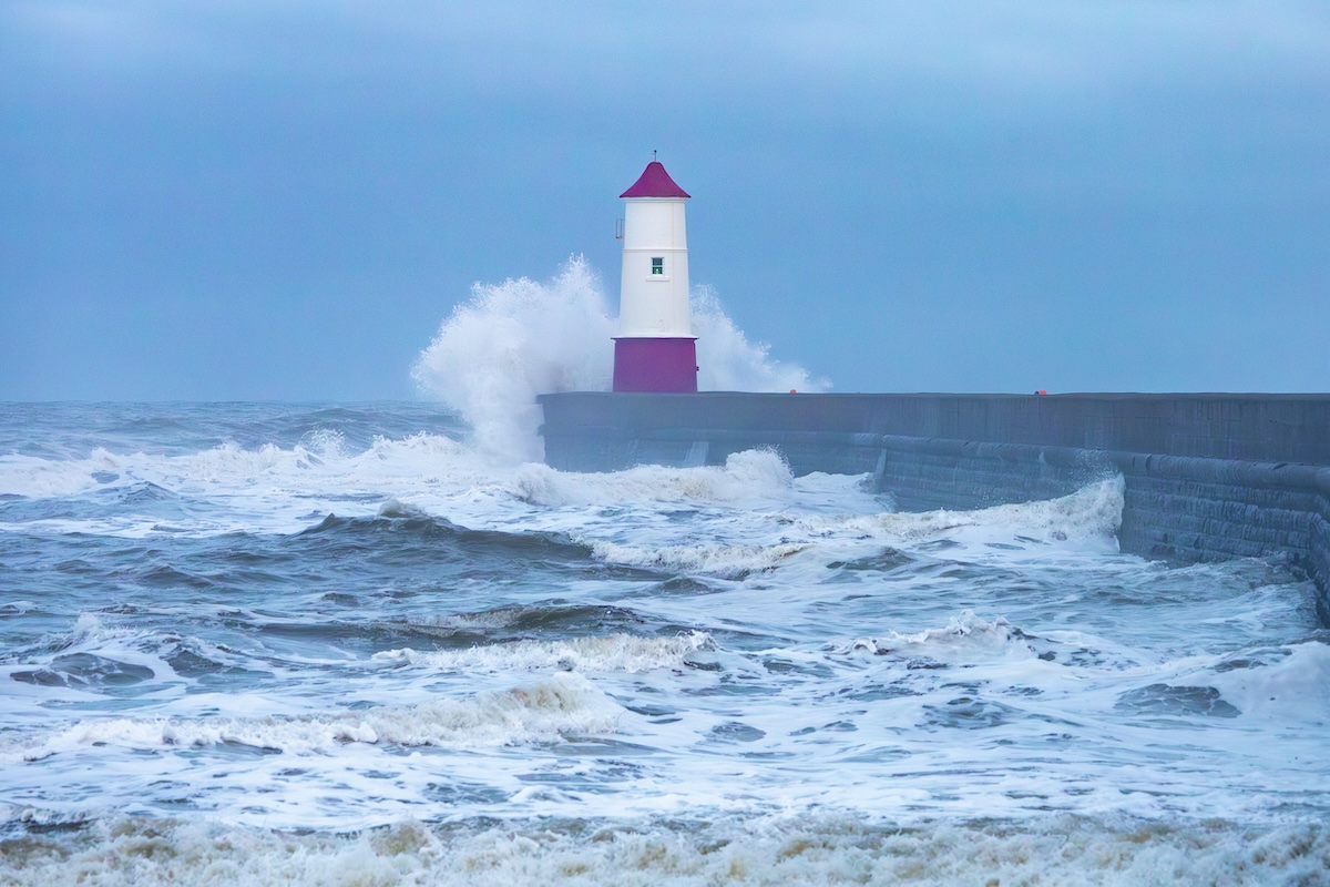 A lighthouse with a red roof stands on a pier as waves crash against it, creating a dramatic spray. The sea is rough, and the sky is overcast, giving the scene a moody atmosphere.