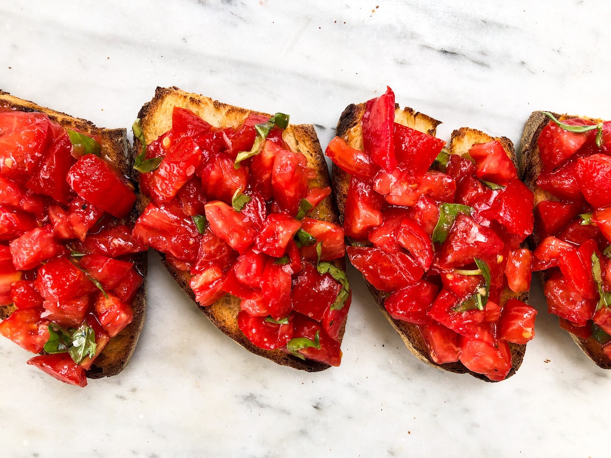 Toasted slices of Italian bread topped with chopped ripe tomatoes, fresh basil, and olive oil—vegan bruschetta served on a marble surface.