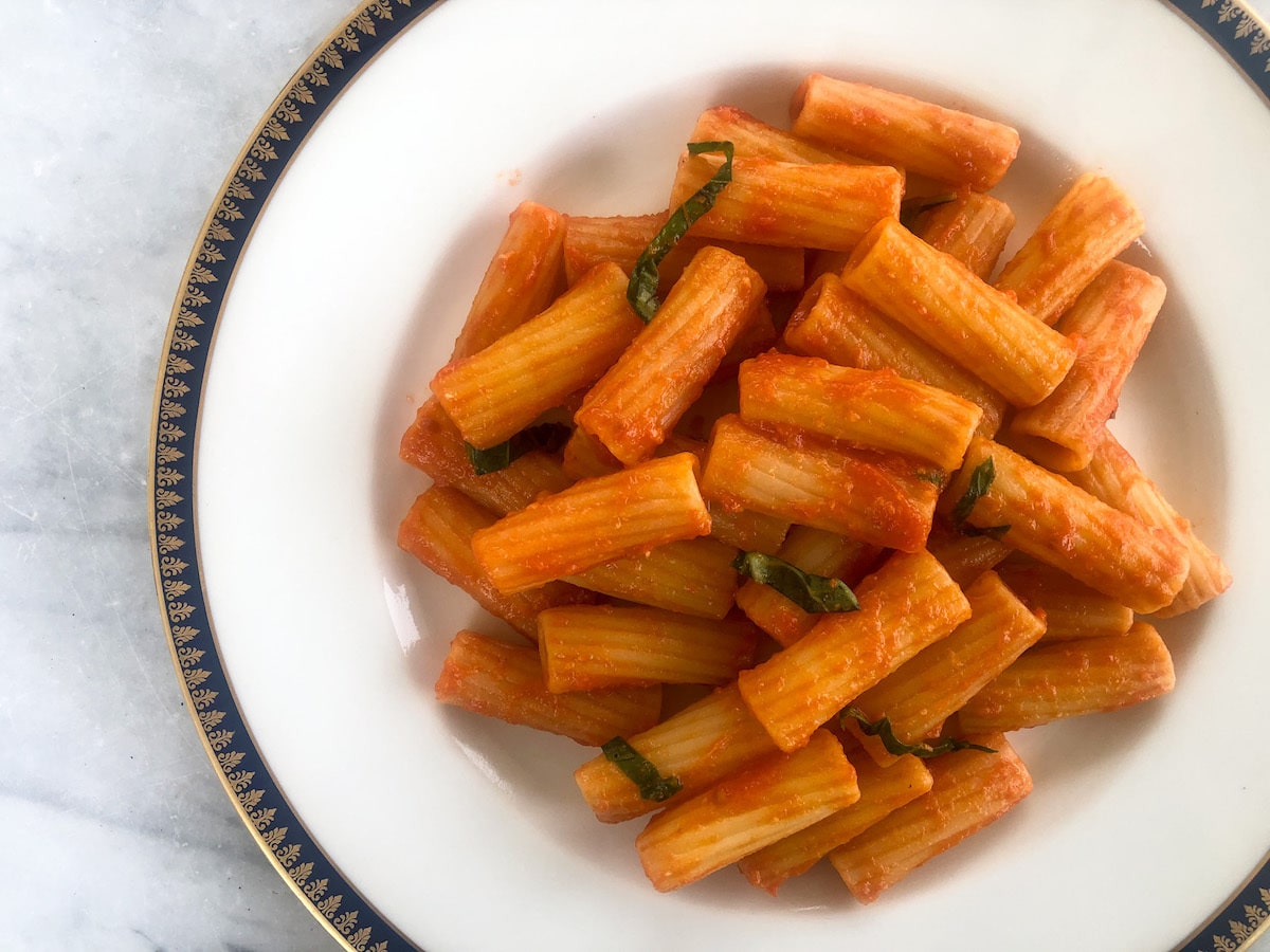A plate of pasta al pomodoro featuring rigatoni tossed in a simple tomato sauce and garnished with fresh basil, served on a white porcelain dish with a decorative blue and gold rim.