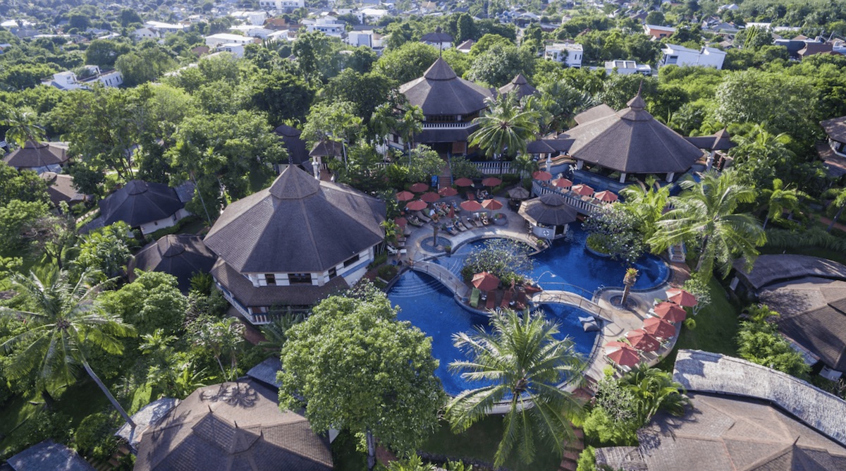 An aerial view of the Mangosteen Ayurveda & Wellness Resort, a vegan-friendly resort in Phuket, Thailand. The resort features multiple thatched-roof villas surrounded by lush tropical greenery. At the center, there's a large, beautifully designed pool area with lounge chairs and umbrellas, creating a serene and luxurious atmosphere for relaxation and wellness activities.