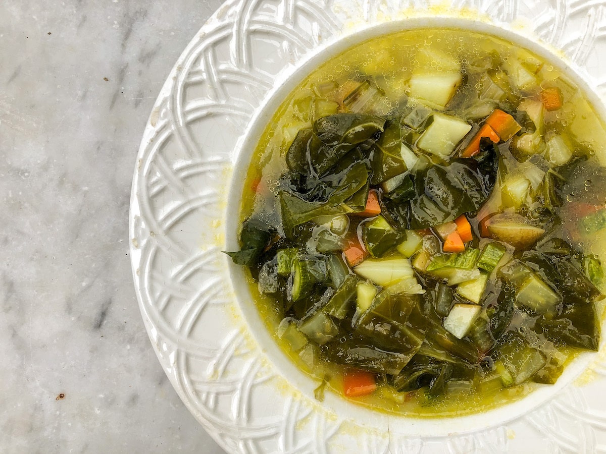 A bowl of seasonal vegan minestrone soup filled with chopped greens, carrots, zucchini, and potatoes in a golden vegetable broth, served on a white ceramic plate.