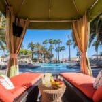 A cozy cabana overlooking the pool at the vegan-friendly Mission Bay Resort in San Diego, California, with vibrant orange cushions, a refreshing fruit platter, and palm trees in the background.