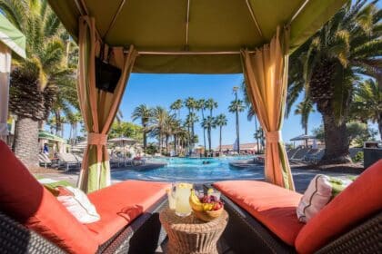 A cozy cabana overlooking the pool at the vegan-friendly Mission Bay Resort in San Diego, California, with vibrant orange cushions, a refreshing fruit platter, and palm trees in the background.
