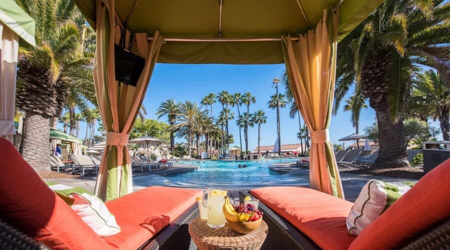 A cozy cabana overlooking the pool at the vegan-friendly Mission Bay Resort in San Diego, California, with vibrant orange cushions, a refreshing fruit platter, and palm trees in the background.